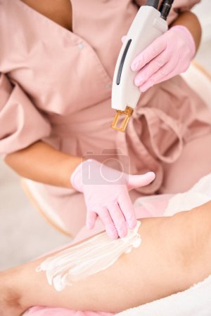 Téléchargez les photos : Cosmetologue lubrifie les mains des patients avec une crème protectrice avant une procédure d'épilation au laser, à l'aide d'un dispositif moderne - en image libre de droit