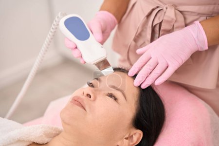 Foto de Hermosa mujer sometida a limpieza facial ultrasónica en un salón de cosmetología, un esteticista utiliza un dispositivo moderno - Imagen libre de derechos
