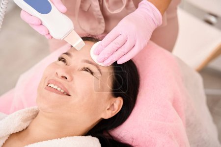Foto de Mujer asiática sometida a limpieza facial ultrasónica en un salón de cosmetología, un especialista utiliza un dispositivo moderno - Imagen libre de derechos