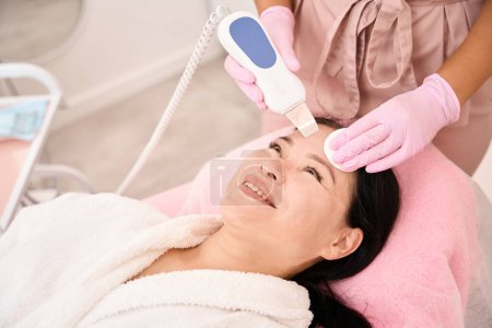Photo for Woman undergoing ultrasonic facial cleansing in a cosmetology salon, a specialist uses a modern device - Royalty Free Image