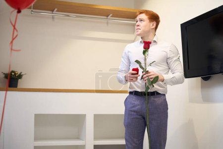 Téléchargez les photos : Jeune homme en chemise blanche et pantalon violet avec rose rouge regardant loin tout en se tenant dans la chambre moderne - en image libre de droit