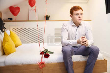 Téléchargez les photos : Homme attrayant assis sur le lit orné de roses rouges et de ballons en forme de coeur dans la chambre d'hôtel - en image libre de droit