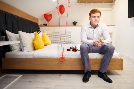 Photo for Fashionable male with red hair sitting amidst red heart balloons and roses in modern bedroom - Royalty Free Image