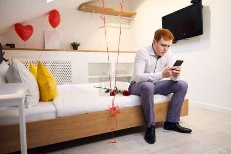 Foto de Hombre pelirrojo sentado en el borde de la cama y el teléfono celular de navegación en el dormitorio moderno - Imagen libre de derechos