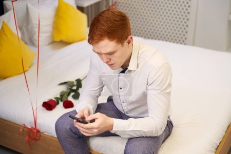 Téléchargez les photos : Jeune homme absorbé dans son téléphone assis sur un lit blanc orné de roses rouges dans la chambre d'hôtel - en image libre de droit