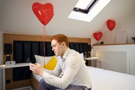Foto de Diligente hombre sentado en la cama con globos rojos y el uso de teléfono celular en el dormitorio moderno - Imagen libre de derechos