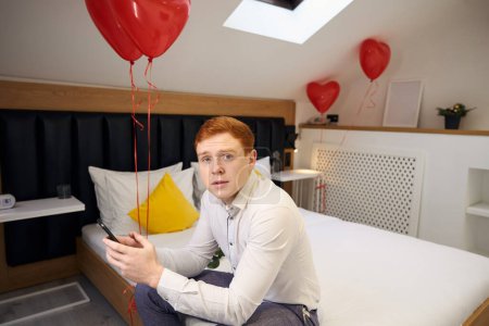 Téléchargez les photos : Jeune homme assis sur le lit avec des ballons rouges et tenant mobile tout en regardant la caméra dans la chambre moderne - en image libre de droit