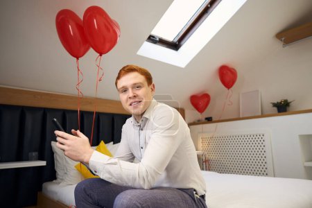 Téléchargez les photos : Homme souriant en chemise blanche assis sur le lit avec des ballons de coeur rouge et tenant le téléphone dans une chambre confortable - en image libre de droit