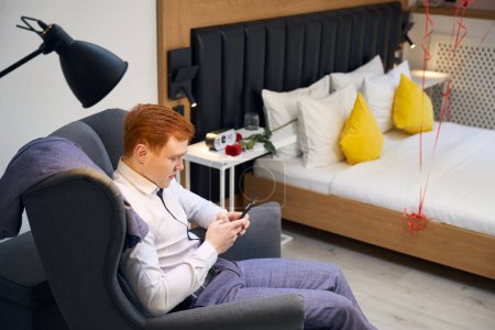 Téléchargez les photos : Homme à la mode assis dans un fauteuil bleu et un téléphone portable de navigation dans un appartement contemporain - en image libre de droit