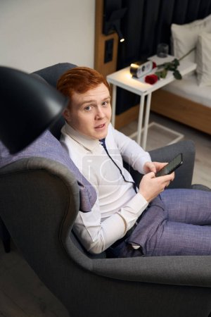 Téléchargez les photos : Vue du dessus de l'homme confiant en costume relaxant dans un fauteuil bleu et tenant le téléphone dans une chambre élégante avec lit - en image libre de droit