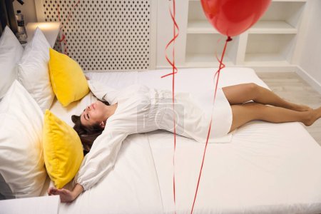 Téléchargez les photos : Belle femme dormant dans un lit blanc avec des ballons rouges et rêvant d'une occasion spéciale avec son partenaire dans la chambre d'hôtel - en image libre de droit