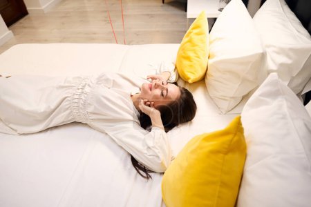 Téléchargez les photos : Femme fatiguée en robe blanche couchée sur un lit confortable avec des oreillers jaunes dans un appartement contemporain - en image libre de droit