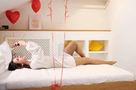 Téléchargez les photos : Femme souriante en robe blanche se détendre sur le lit avec des ballons coeur rouge dans un appartement contemporain - en image libre de droit