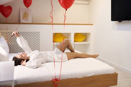 Téléchargez les photos : Jeune femme en robe blanche se prélasser sur le lit et tenant mobile avec des ballons coeur rouge dans la chambre moderne - en image libre de droit