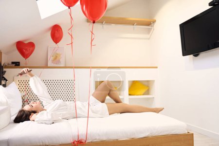 Téléchargez les photos : Jeune femme en robe blanche couchée sur le lit avec des ballons coeur rouge dans une chambre lumineuse et spacieuse - en image libre de droit
