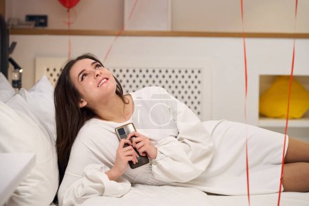 Téléchargez les photos : Femme souriante en robe blanche tenant son téléphone et regardant le plafond tout en étant couché sur un lit blanc dans la chambre d'hôtel - en image libre de droit