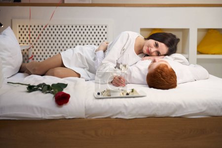 Téléchargez les photos : Aimant homme et femme profitant d'un repas romantique au lit avec rose rouge et plateau de dessert dans la chambre d'hôtel - en image libre de droit
