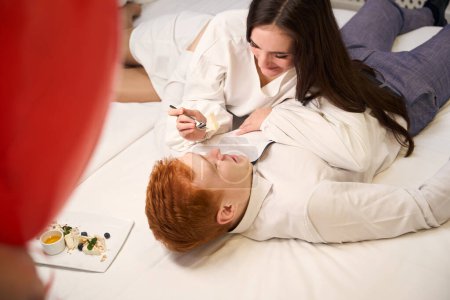 Téléchargez les photos : Vue du dessus de la femme attrayante en robe blanche nourrissant son petit ami tout en étant couché sur le lit dans la chambre d'hôtel - en image libre de droit