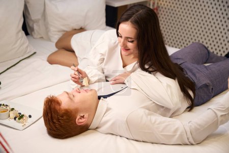 Téléchargez les photos : Belle femme en robe blanche nourrissant son petit ami tout allongé sur le lit dans la chambre d'hôtel - en image libre de droit