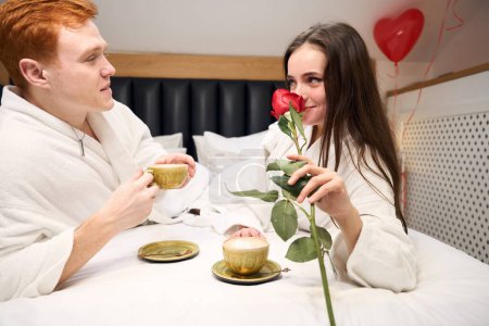 Téléchargez les photos : Aimant homme et femme en robe blanche profitant d'un petit déjeuner romantique au lit avec café et rose dans la chambre d'hôtel - en image libre de droit