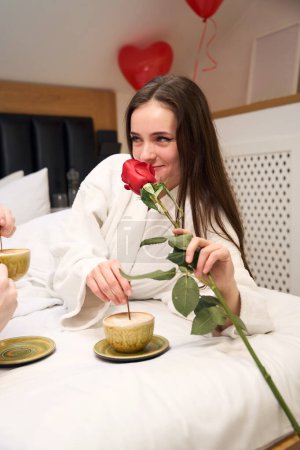 Téléchargez les photos : Belle femme célébrant la Saint-Valentin avec une tasse de café et rose dans une chambre confortable avec ballons coeur rouge - en image libre de droit