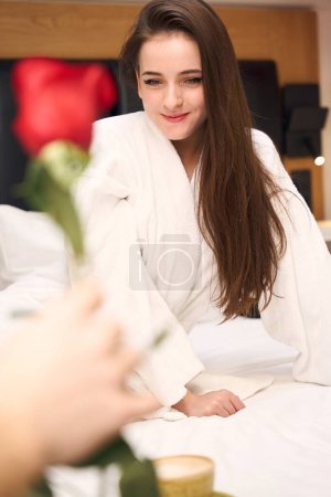 Téléchargez les photos : Femme heureuse en robe blanche assise sur le lit avec rose et tasse de café à proximité dans la chambre d'hôtel - en image libre de droit
