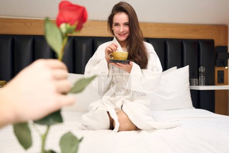 Téléchargez les photos : Femme souriante en robe blanche tenant tasse de café et assis sur le lit est offert rose à la main de l'homme à l'intérieur - en image libre de droit