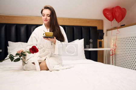 Téléchargez les photos : Femme heureuse profitant d'une tasse de café et rose sur un lit confortable avec des ballons de coeur dans un appartement contemporain - en image libre de droit