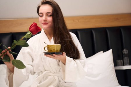 Téléchargez les photos : Belle femme en robe blanche profitant d'une tasse de café et rose tout en étant assis sur un lit confortable dans un appartement contemporain - en image libre de droit