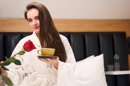 Téléchargez les photos : Jolie femme en robe blanche profitant d'une tasse de café et rose tout en étant assis sur un lit confortable dans un appartement contemporain - en image libre de droit