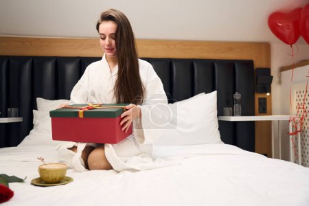 Téléchargez les photos : Jolie femme en robe blanche assise sur le lit et tenant boîte cadeau rouge dans un appartement contemporain - en image libre de droit