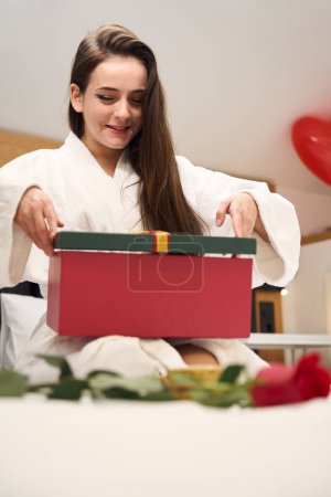 Téléchargez les photos : Joyeuse femelle en robe blanche assise sur le lit et ouvrant boîte cadeau rouge dans un appartement contemporain - en image libre de droit