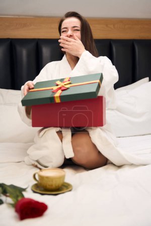 Foto de Mujer feliz en bata blanca sentada en la cama y bostezando mientras se abre presente en la habitación del hotel - Imagen libre de derechos