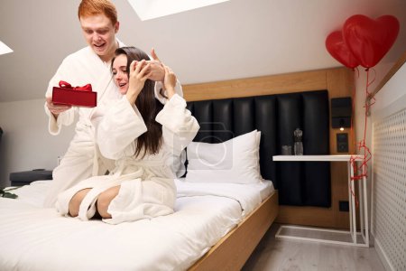 Téléchargez les photos : Femme surprise assise sur le lit comme son partenaire donnant sa boîte-cadeau rouge dans des appartements contemporains - en image libre de droit