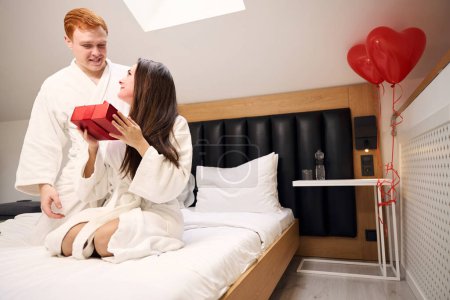 Foto de Atractiva hembra en bata blanca recibiendo un regalo de su novio en un apartamento contemporáneo - Imagen libre de derechos