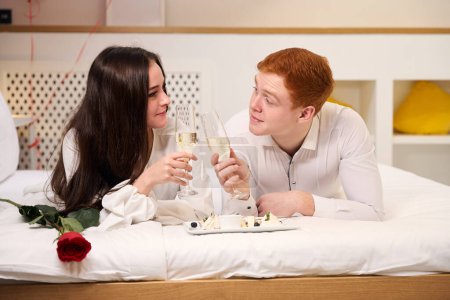 Photo for Happy couple enjoying romantic moment with champagne and dessert while lying on bed in contemporary apartment - Royalty Free Image
