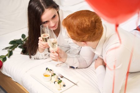 Téléchargez les photos : Vue de dessus du jeune homme et de la jeune femme se regardant et cliquetant des verres de champagne au lit - en image libre de droit