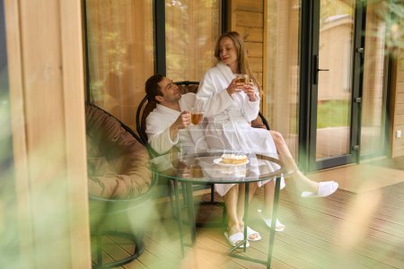 Téléchargez les photos : Deux jeunes amants en peignoirs blancs passent le week-end ensemble assis sur une chaise et boivent des boissons sur le porche - en image libre de droit