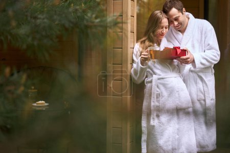 Téléchargez les photos : Jolie femme en peignoir blanc buvant du thé et recevant un cadeau boîte rouge de son doux petit ami, debout sur le porche - en image libre de droit