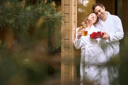 Téléchargez les photos : Jolie femme en peignoir blanc buvant du thé et recevant un cadeau boîte rouge de son homme doux, debout sur le porche - en image libre de droit