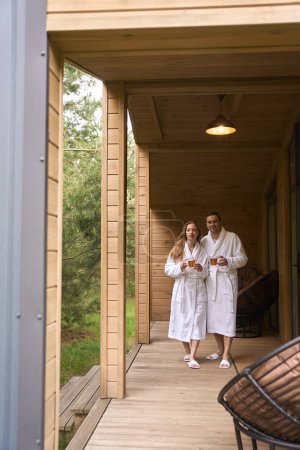 Photo for Handsome man and beautiful woman in white bathrobes embracing and drinking fresh relaxing tea - Royalty Free Image
