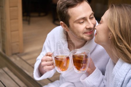 Photo for Romantic couple in robes rubbing noses as sign of love while sitting on doorstep and drinking morning tea - Royalty Free Image