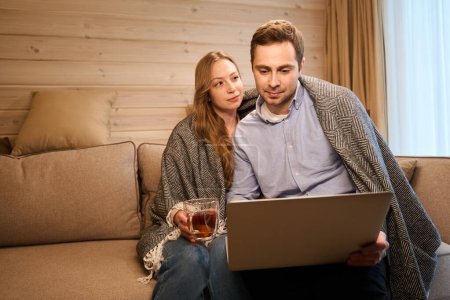 Téléchargez les photos : Homme utilisant ordinateur portable assis détendu sur le canapé sous plaid tandis que la petite amie boire du thé et le regarder - en image libre de droit