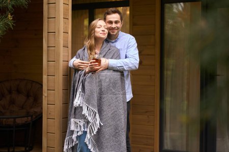 Photo for Happy young male embracing his pretty lady wrapped in blanket and drinking morning beverage while they standing on porch - Royalty Free Image
