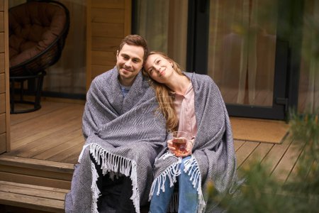 Foto de Mujer sonriente bebiendo té caliente apoyada en su marido. Dulce pareja cubierta de mantas salón en el porche - Imagen libre de derechos