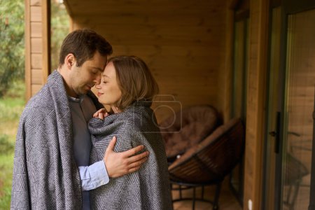 Foto de Jóvenes amantes lindos cubiertos de manta abrazando y disfrutando de momento íntimo preparándose para besarse con los ojos cerrados - Imagen libre de derechos