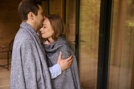 Foto de Marido romántico abrazos y besos en la frente su joven esposa de pie en el porche con los ojos cerrados - Imagen libre de derechos