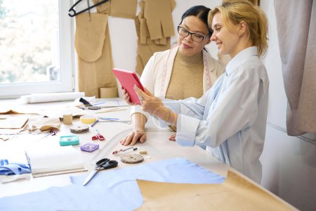 Foto de Las diseñadoras de moda utilizan una tableta en un entorno de trabajo, hay un montón de suministros de costura en la mesa - Imagen libre de derechos