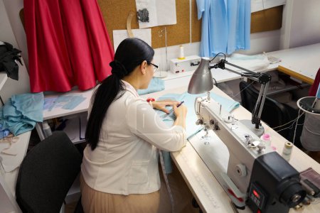 Téléchargez les photos : Dressmaker au travail fonctionne avec du tissu bleu, maître utilise une machine à coudre moderne - en image libre de droit
