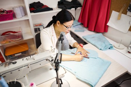 Téléchargez les photos : Maître de couture sur le lieu de travail travaille avec du tissu bleu, la femme utilise une machine à coudre moderne - en image libre de droit
